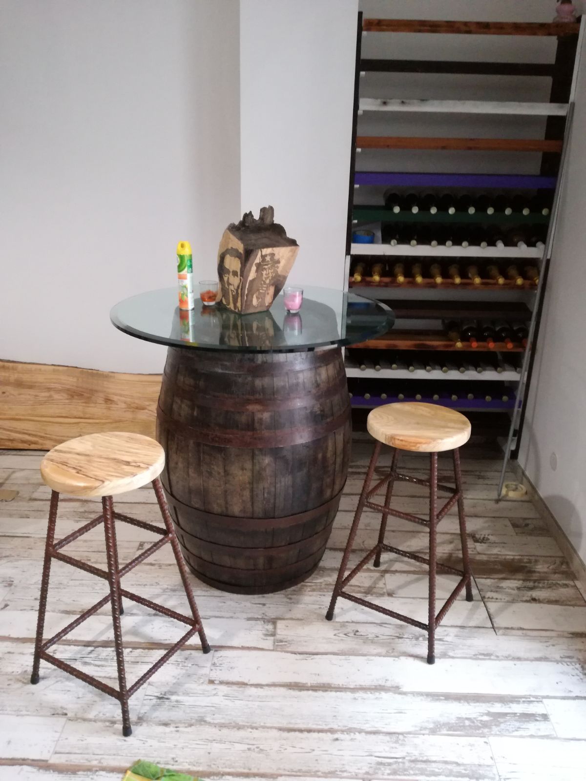 Un angolo bar rustico con un tavolo ricavato da una botte di legno, piano in vetro e due sgabelli, in una stanza spoglia.