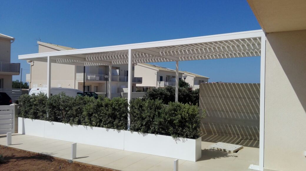 Una moderna pergola bianca sopra un patio delimitato da siepi, accanto a edifici bianchi, sotto un cielo azzurro limpido.