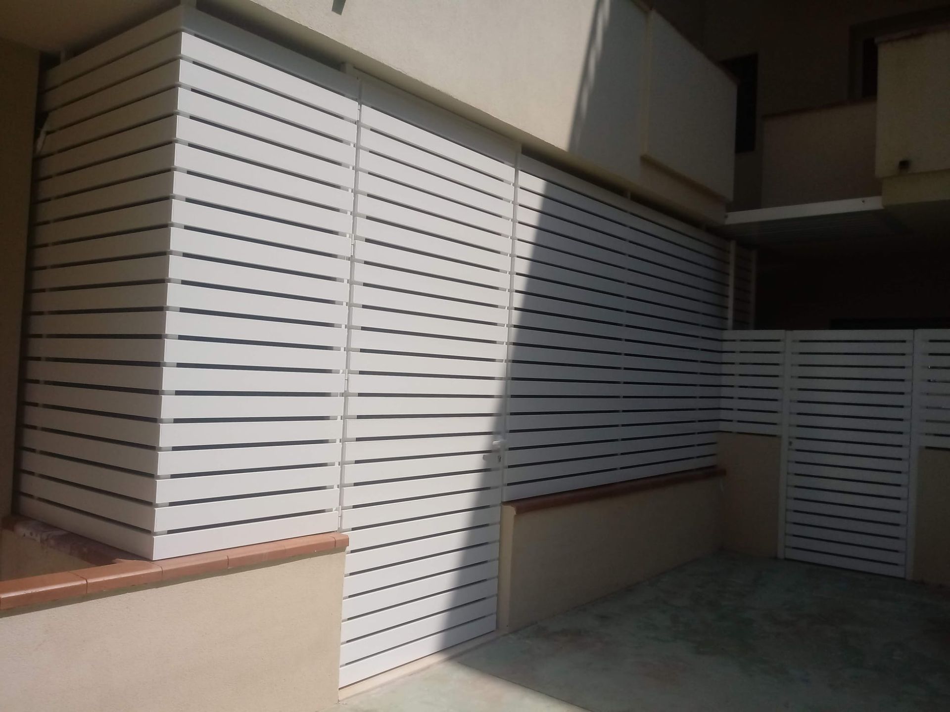 Schermature perimetrali a lamelle bianche sulla facciata di un edificio illuminato dal sole, con un'ombra che si proietta sul muro.