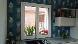 Piccola cucina con carta da parati a motivi blu, finestra, piano di lavoro e mobile bianchi; piante e fiori fuori dalla finestra.
