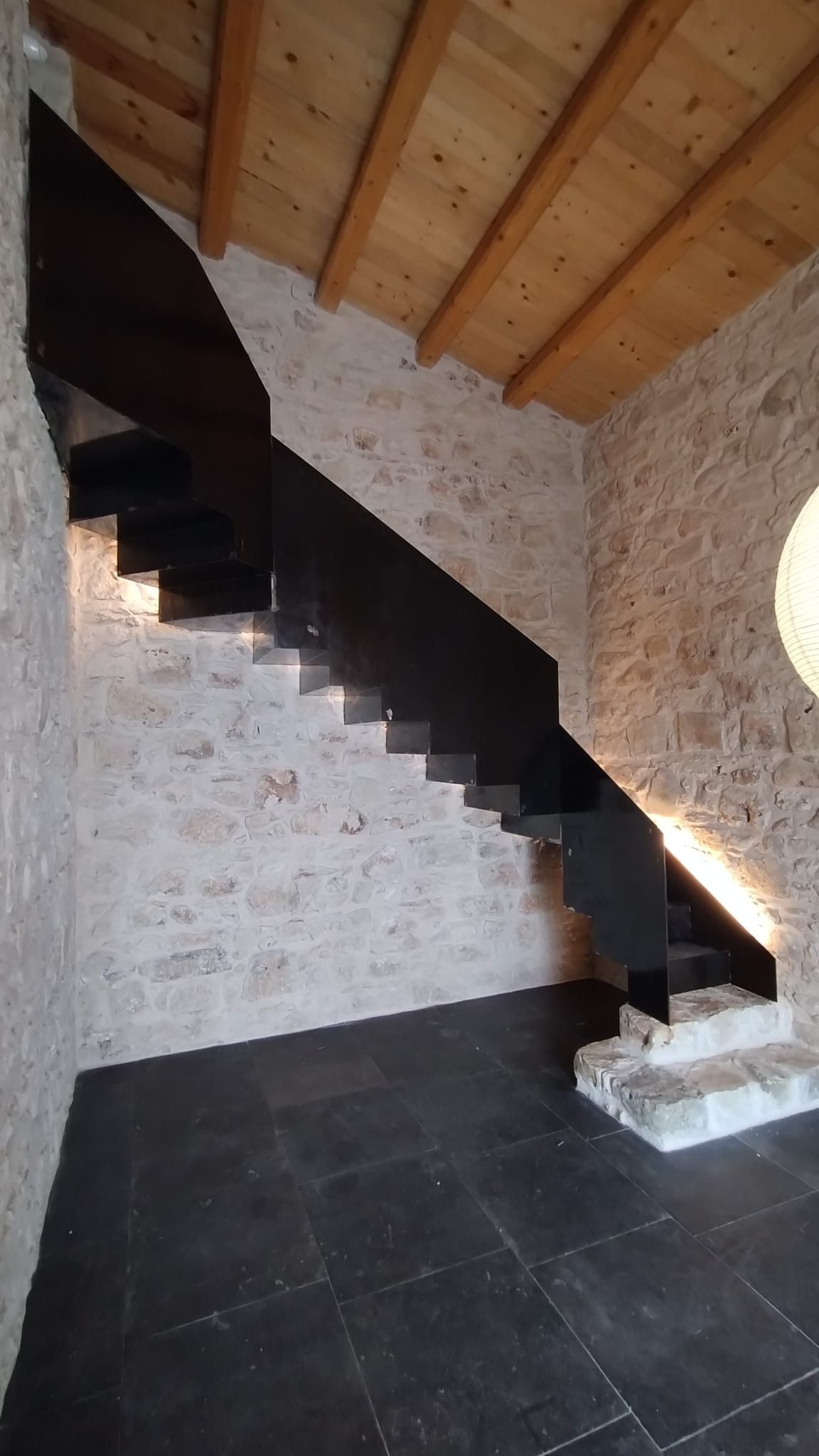 Vano scale in pietra con gradini in legno scuro, pareti bianche con finitura a rilievo e soffitto inclinato con travi in legno.