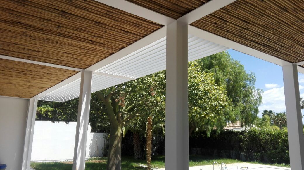 Patio coperto con tetto a doghe di legno, colonne bianche e alberi in un cortile soleggiato