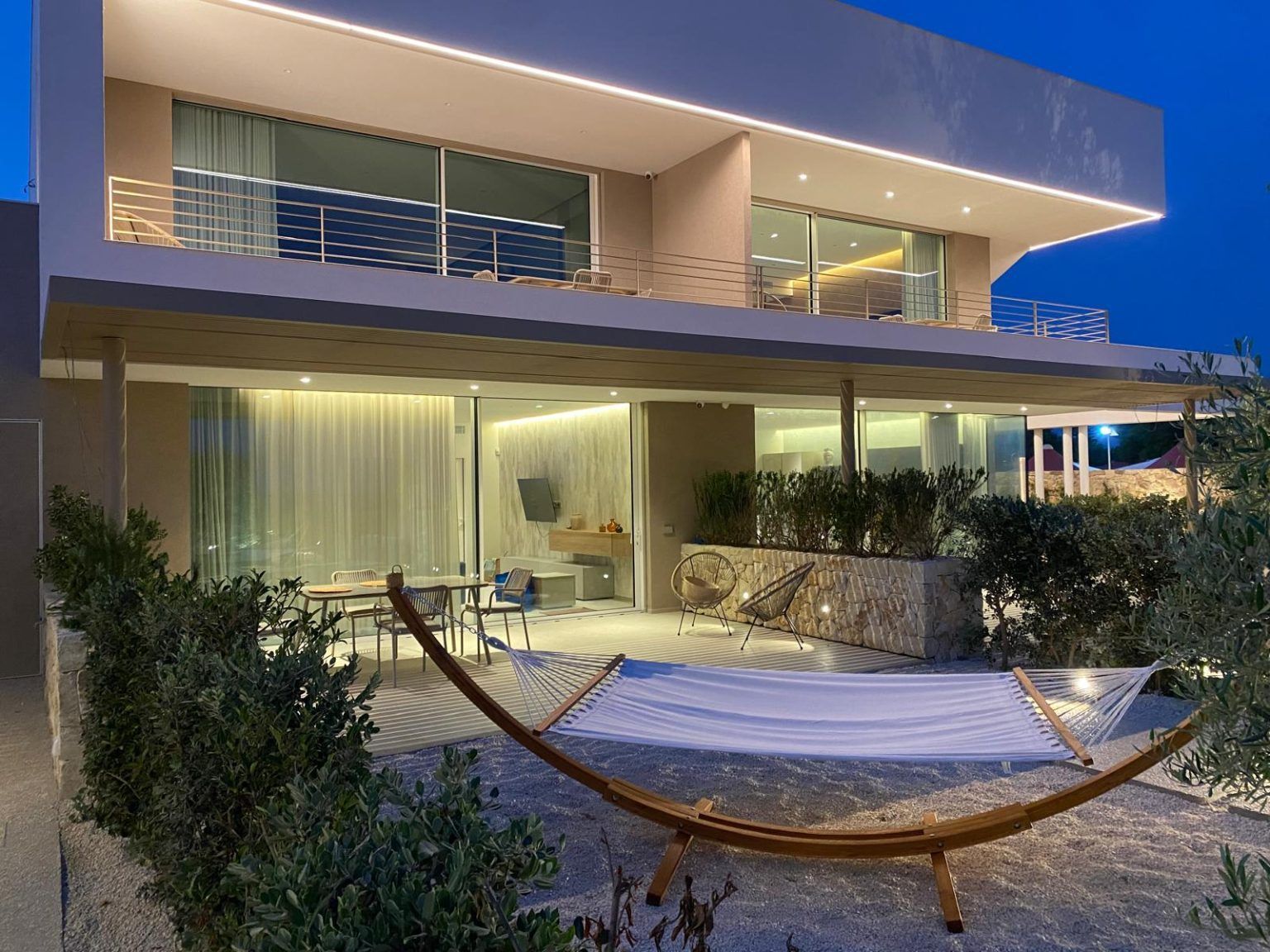 Patio di una casa moderna al crepuscolo con un'amaca sulla ghiaia e grandi porte a vetri illuminate dall'interno