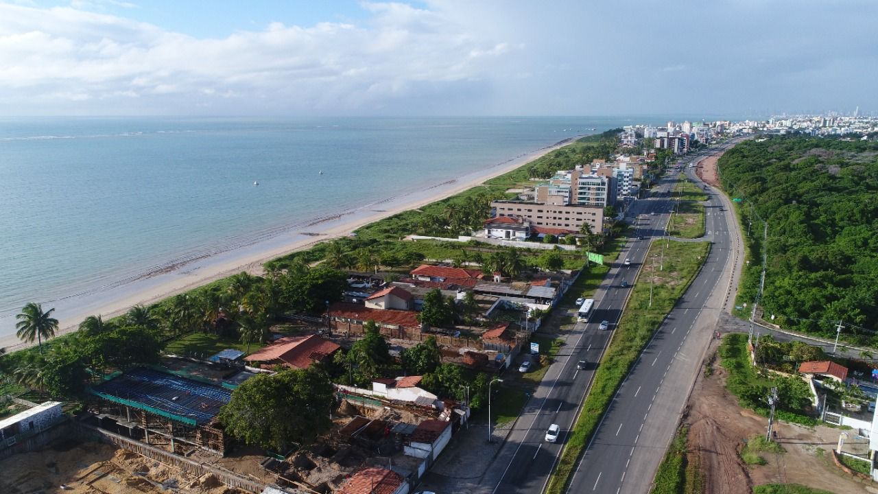 Vista aérea de uma estrada costeira ao lado de uma praia, oceano e encosta verde com casas e edifícios.