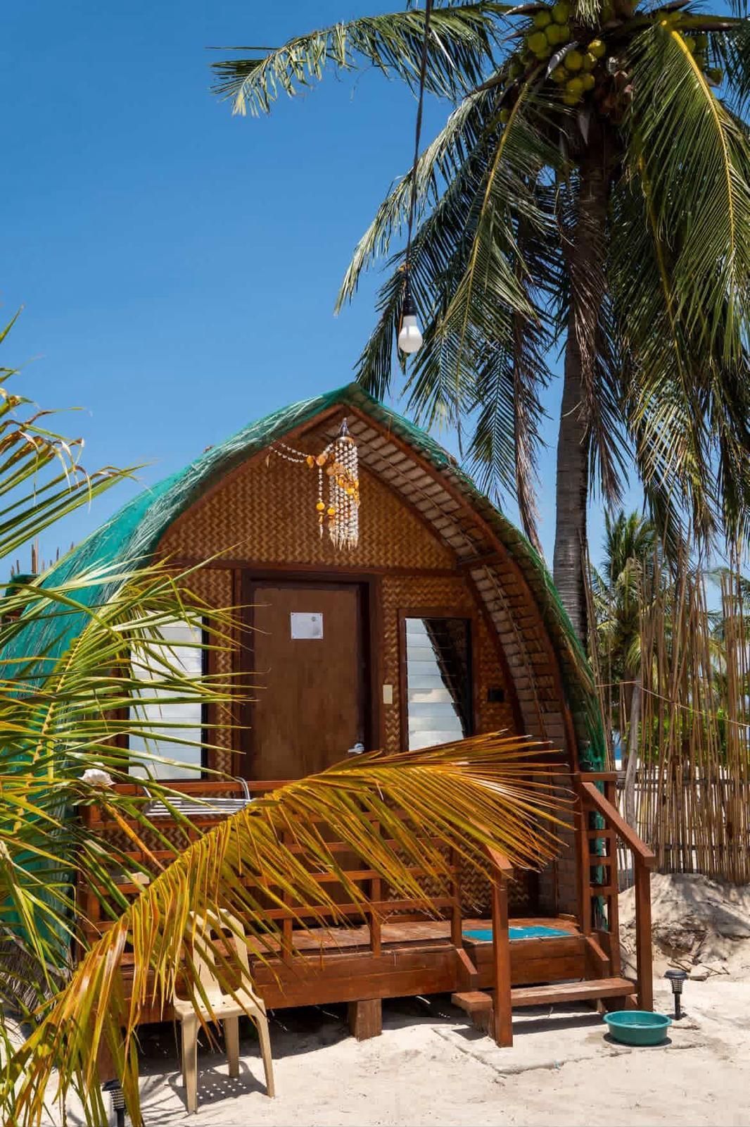 Small bamboo beach hut with a wooden porch under palm trees, against a bright blue sky