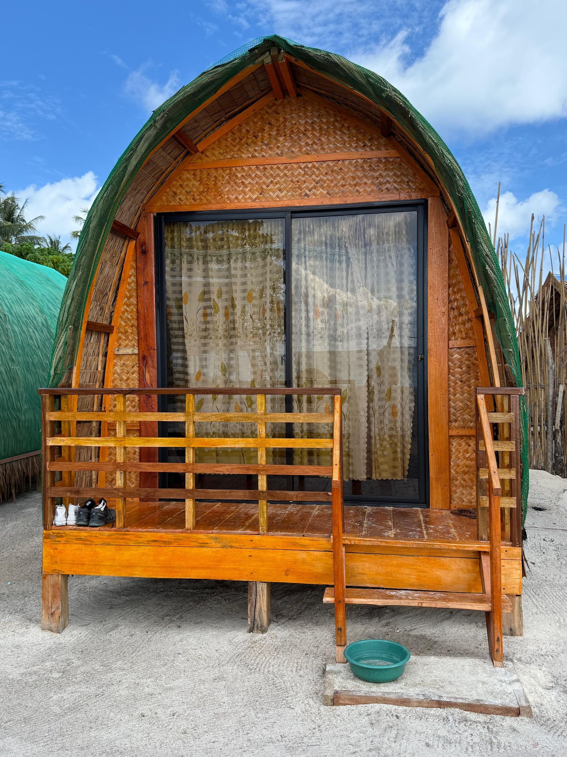 Small wooden cabin with a curved roof and front porch on gravel, with sliding glass doors and green mesh fencing.