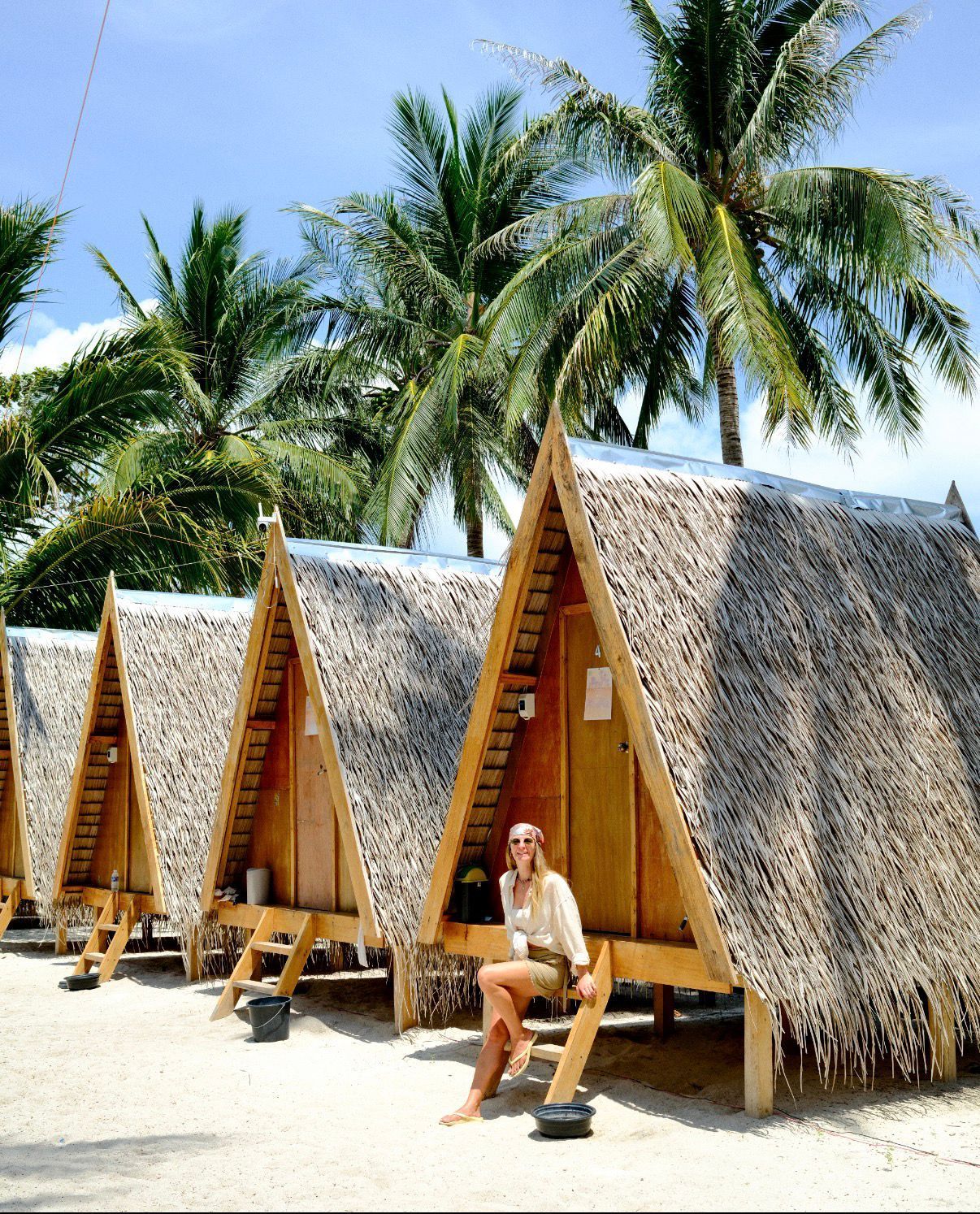 Row of thatched beach huts under palm trees, with a person sitting in front on a sunny beach.