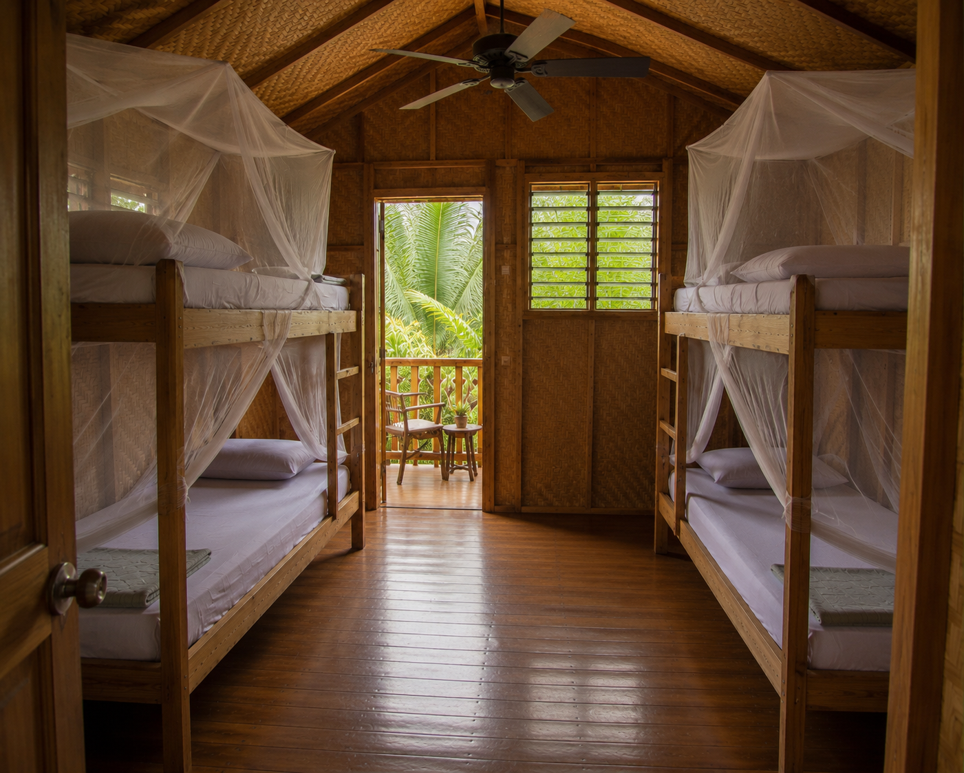 Wooden bunk beds with white mosquito nets in a rustic lodge room, facing a bright doorway to greenery