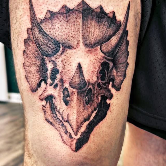 A black and grey tattoo of a Triceratops skull on a person's upper leg.