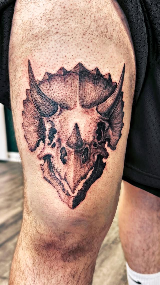 A detailed, black-and-grey tattoo of a triceratops skull on a person's upper thigh.