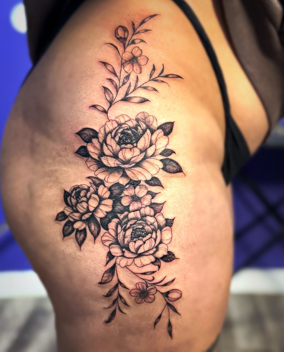 A black and grey floral tattoo featuring multiple peonies and leafy vines extending vertically along a person's thigh.