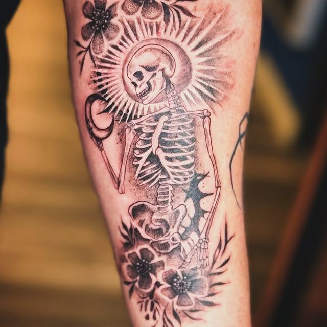 A black and grey tattoo on an arm featuring a skeleton framed by a sunburst, thorns, and floral accents.