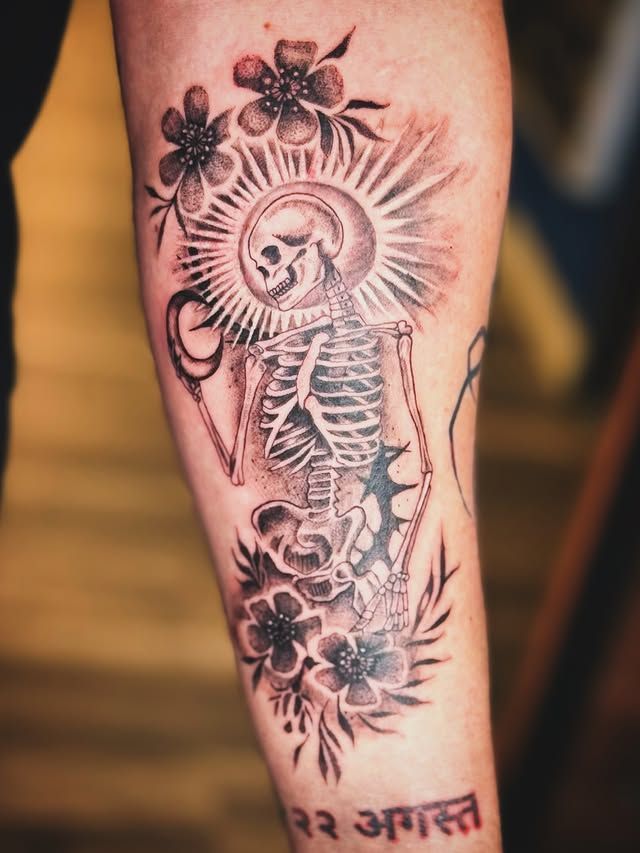 A black-and-grey tattoo of a skeleton holding a mirror, surrounded by flowers and a sunburst, with Hindi text below.