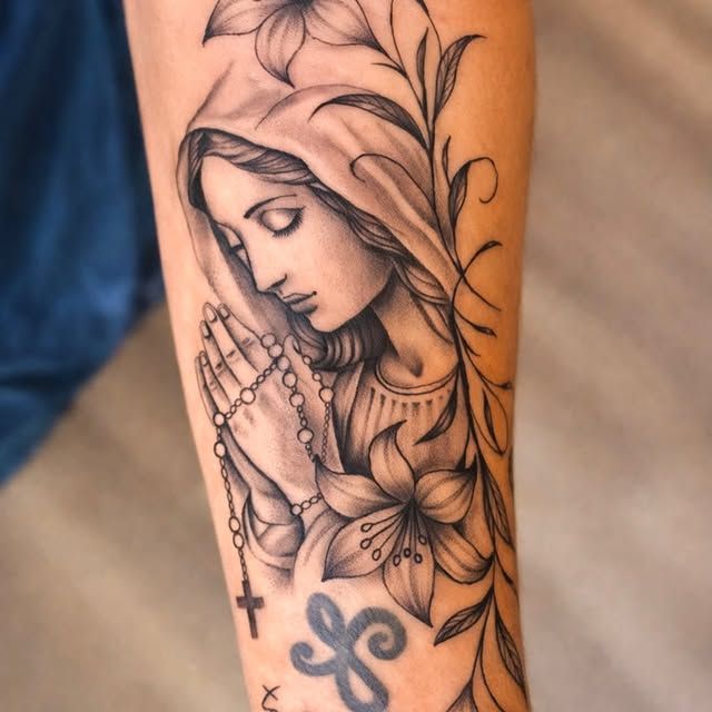 Black-and-grey tattoo of a veiled figure in prayer with a rosary, framed by floral details on a forearm.