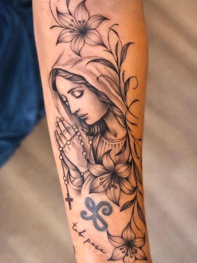 A black-and-grey tattoo of a person in a veil with hands clasped in prayer, surrounded by lilies and a small symbol.