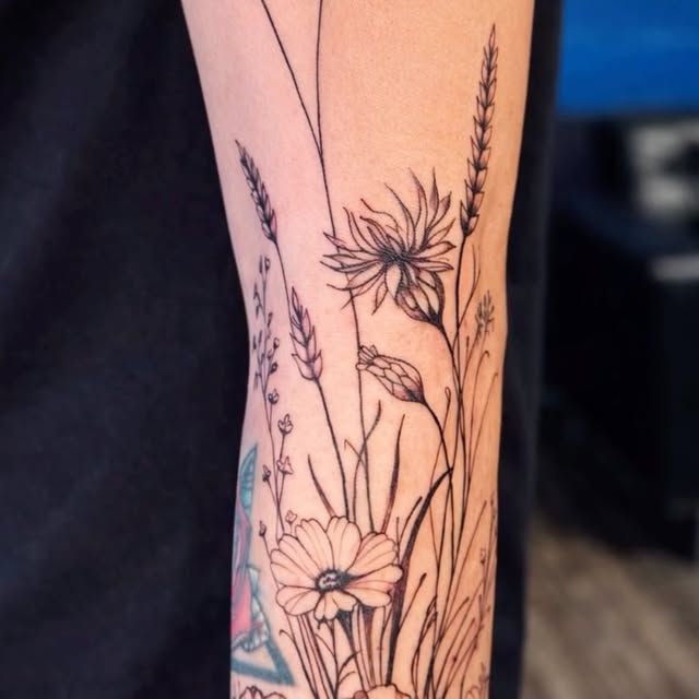 A black-line tattoo on an arm featuring a wildflower bouquet with grasses, stalks, and various delicate blossoms.