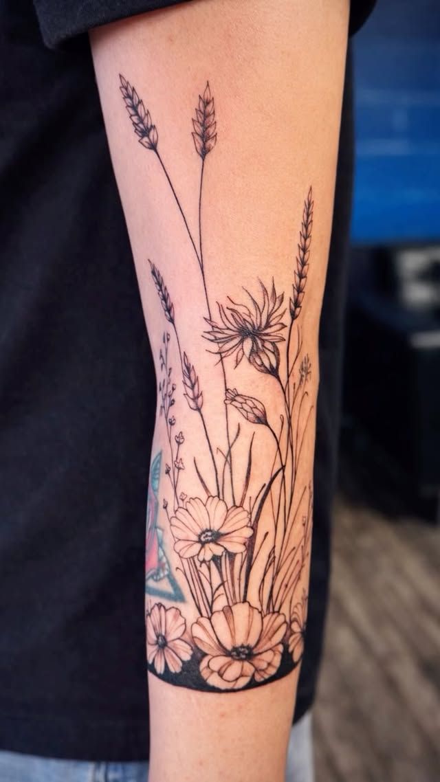 A black-ink tattoo on an arm featuring a botanical arrangement of wildflowers, grasses, and grain stalks.