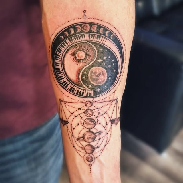 A forearm tattoo featuring a moon phase-crowned yin-yang symbol with a sun and stars, above a geometric planetary alignment.