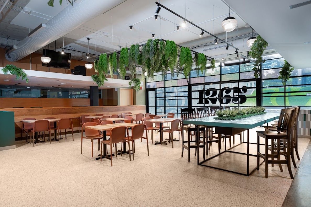 Restaurant interior with tables, chairs, a raised stage, and hanging greenery. Natural light floods the space through large windows.