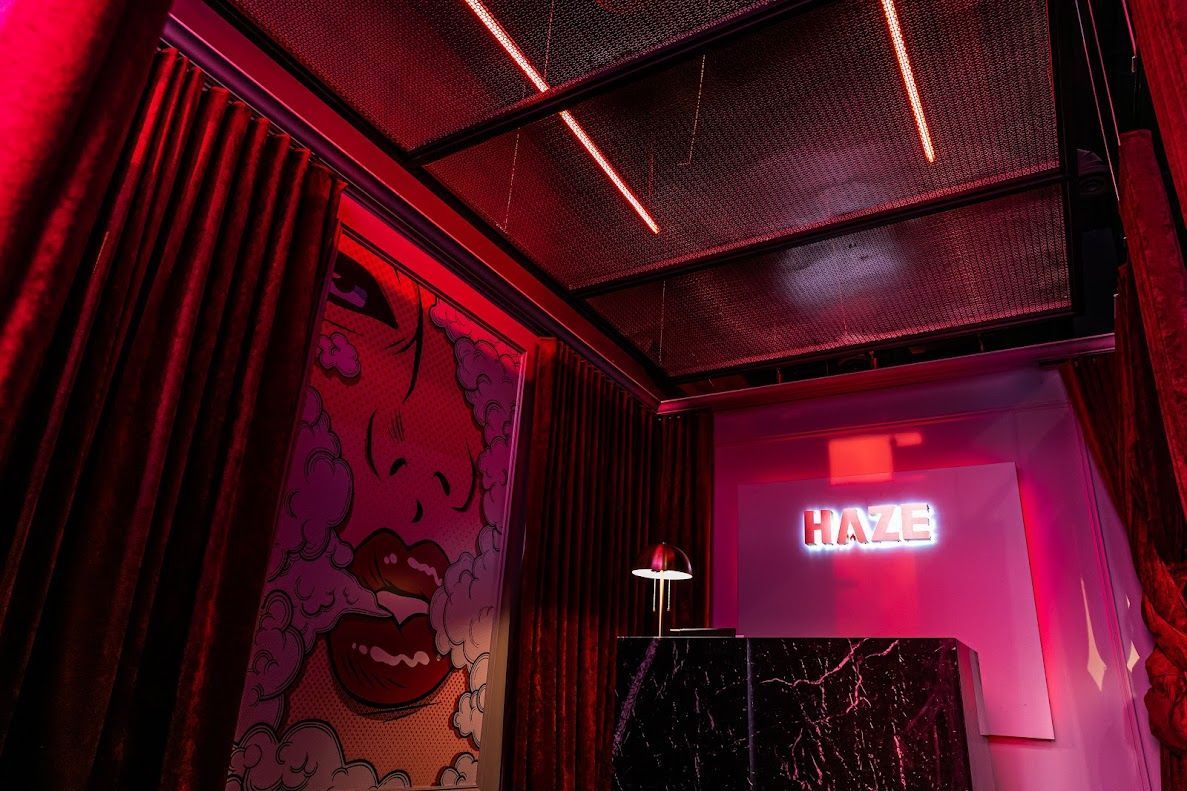 A dimly lit nightclub entrance with red accents. Features include a graffiti-style mural, a marble front desk, and a neon "HAZE" sign.