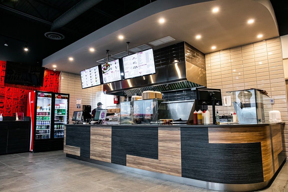 Interior of a restaurant with a counter, menu boards, and a stainless steel kitchen area. Beverage and food displays are visible.