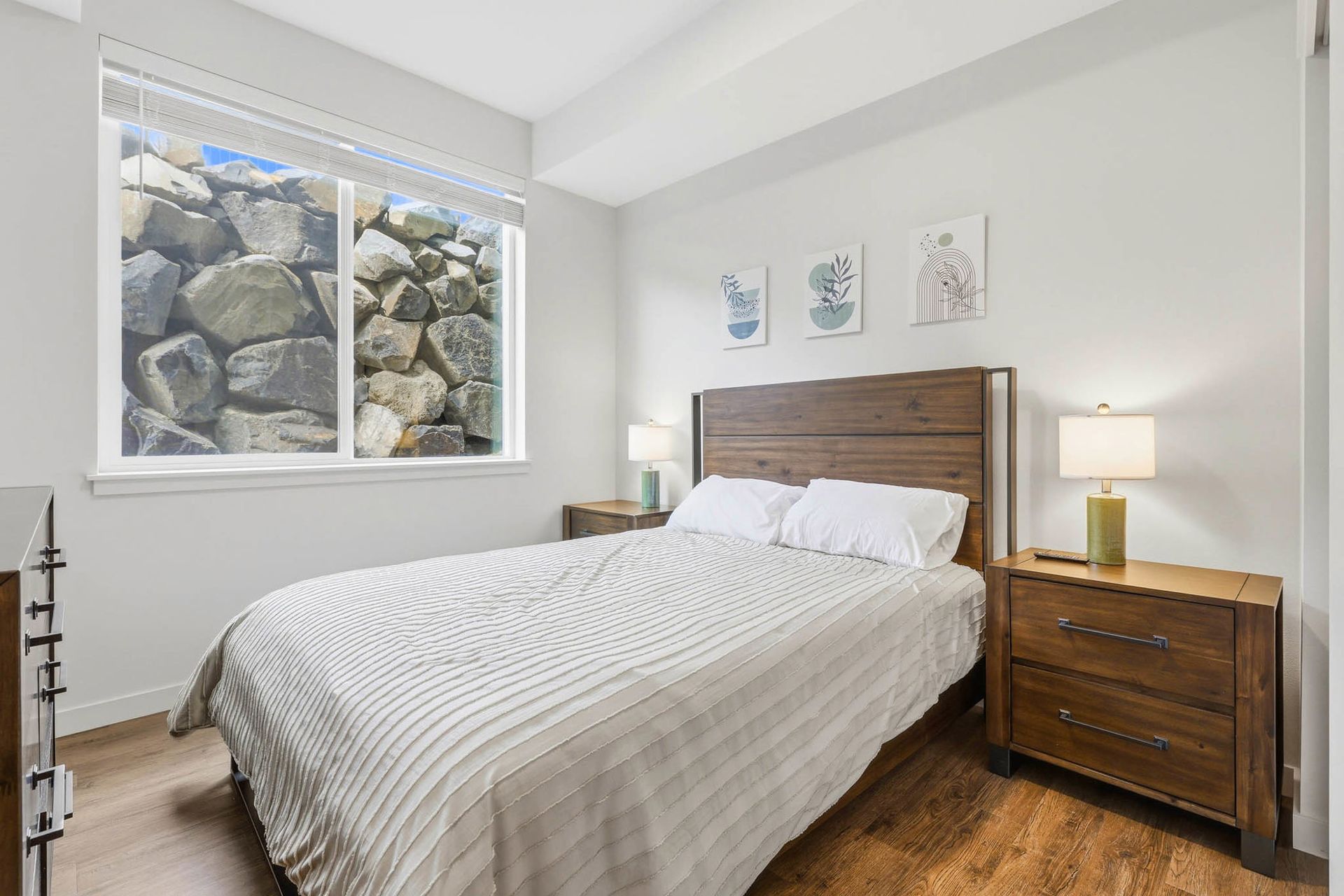 Bright bedroom with a wood bed, nightstands, lamp, hardwood floor, and a window overlooking rocks outside.