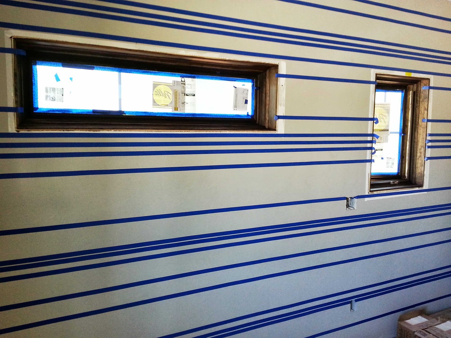 A wall under construction with horizontal blue painter's tape stripes and two window frames installed.