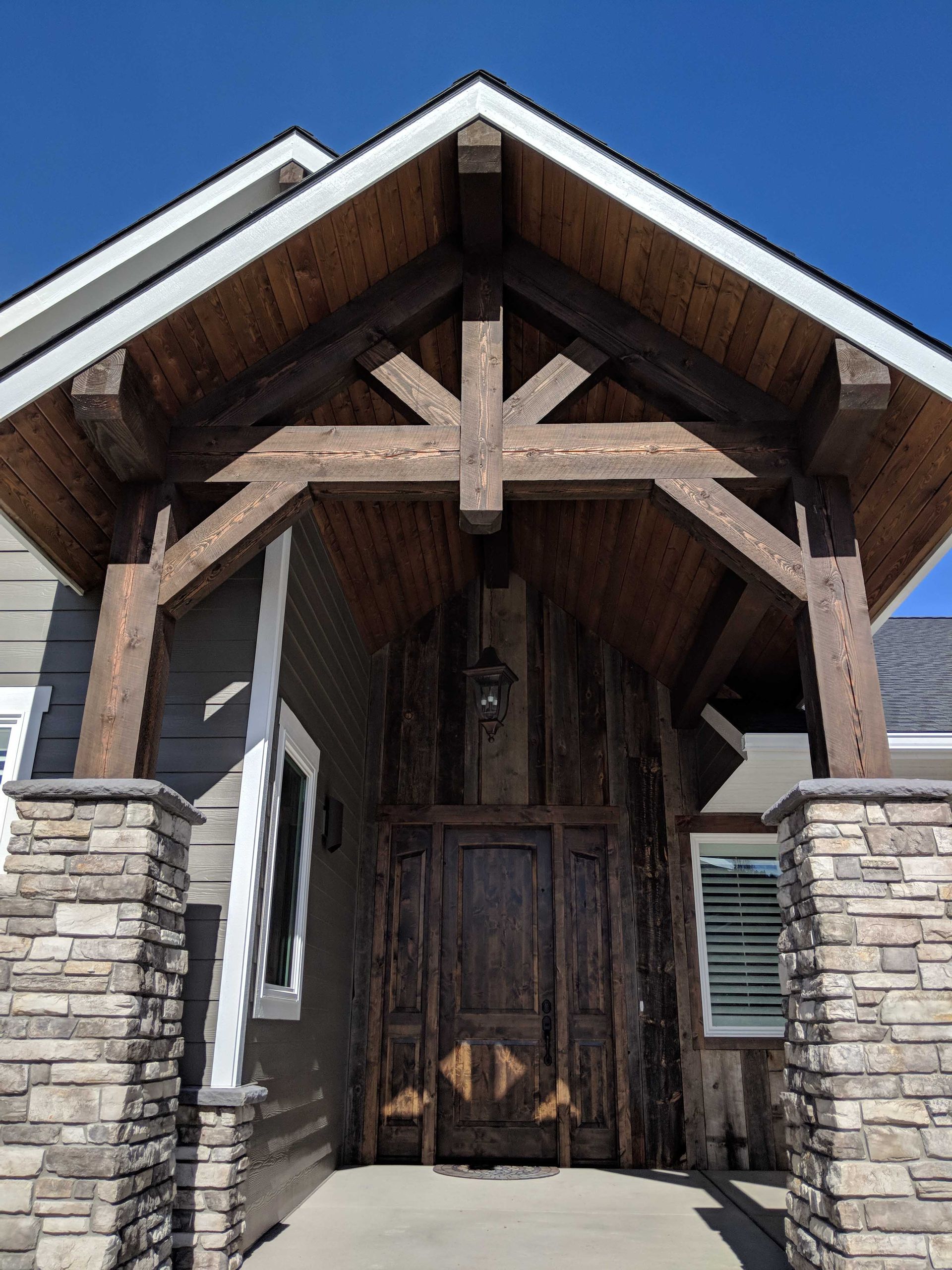 A grand entryway featuring a wooden truss gable, stone pillars, and a decorative dark wood door under a clear blue sky.