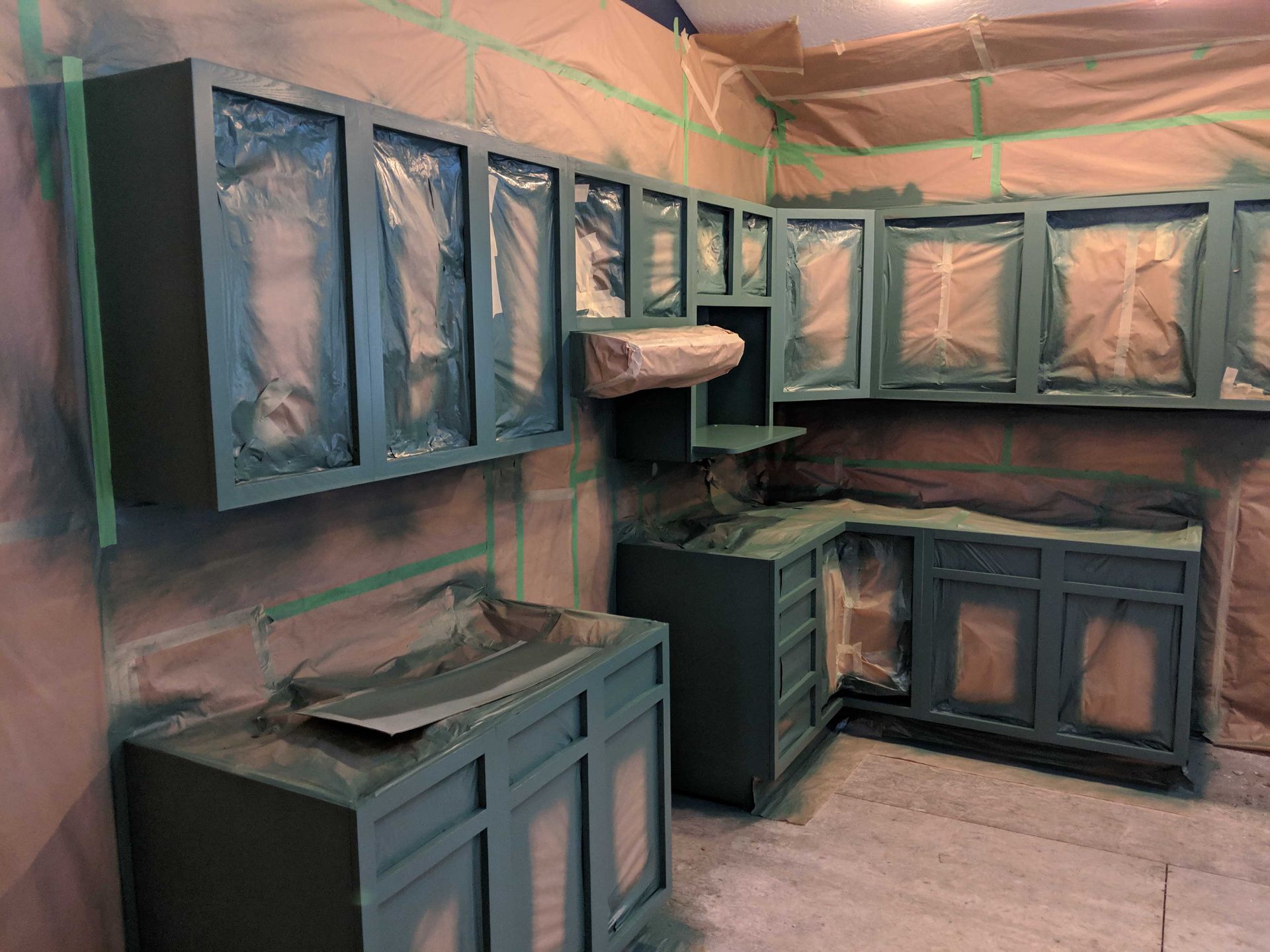 Kitchen cabinets in the process of being painted a dark green, with surfaces masked off with paper and blue tape.