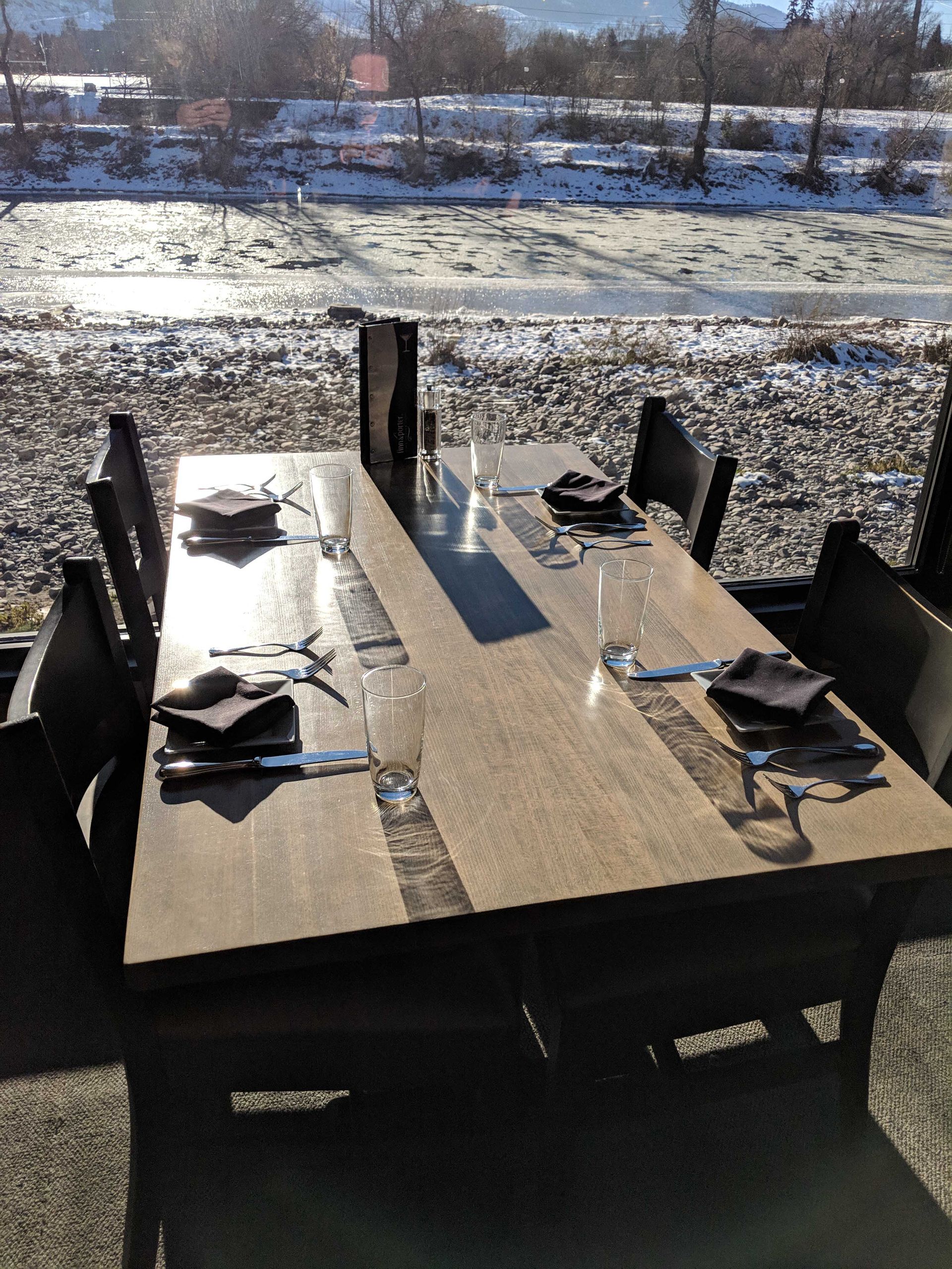 A restaurant table set for four people, overlooking a snowy, icy river on a sunny day.