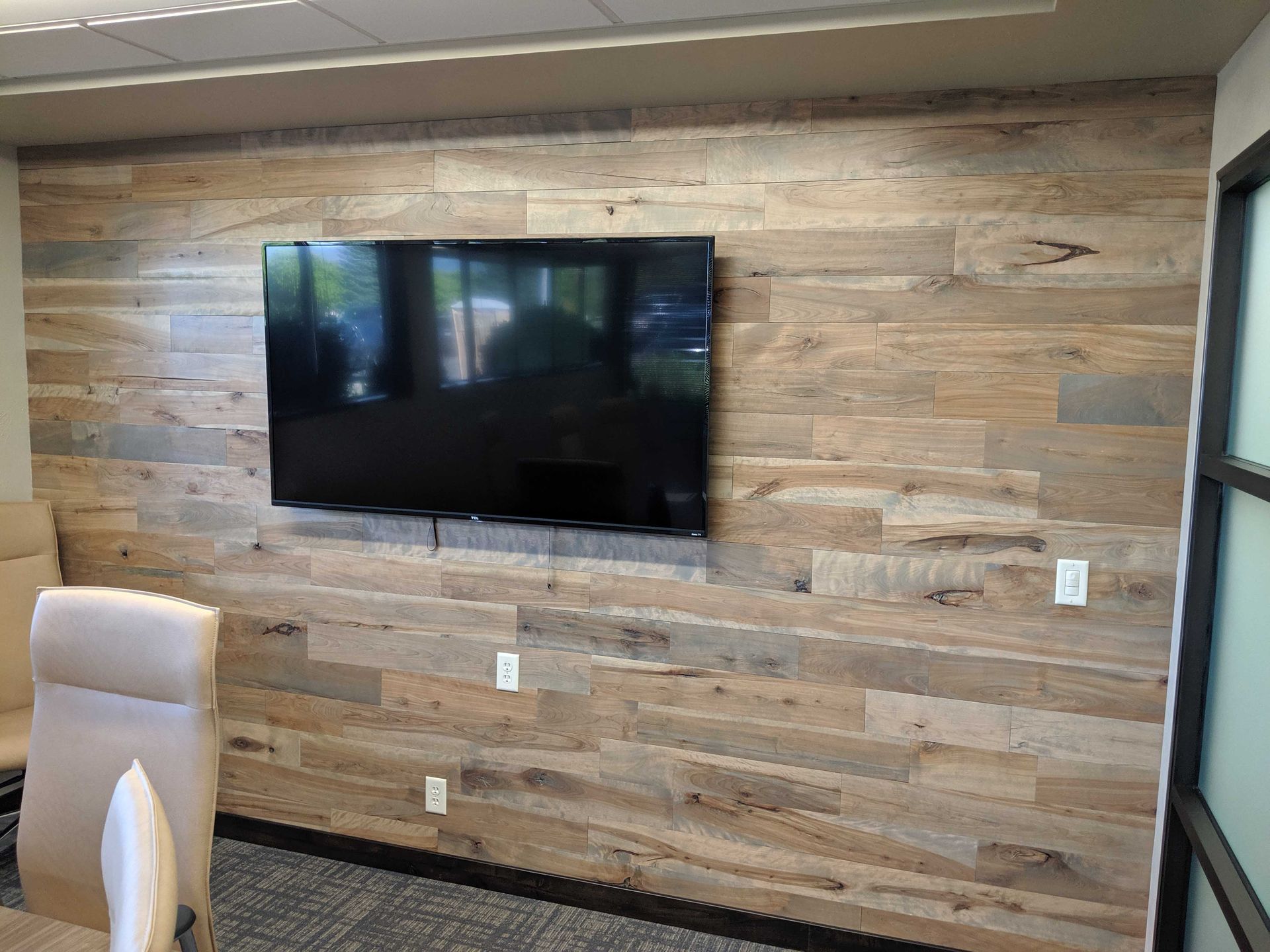 A flat-screen television mounted on a wall covered in horizontal, light-brown wood planks inside an office setting.