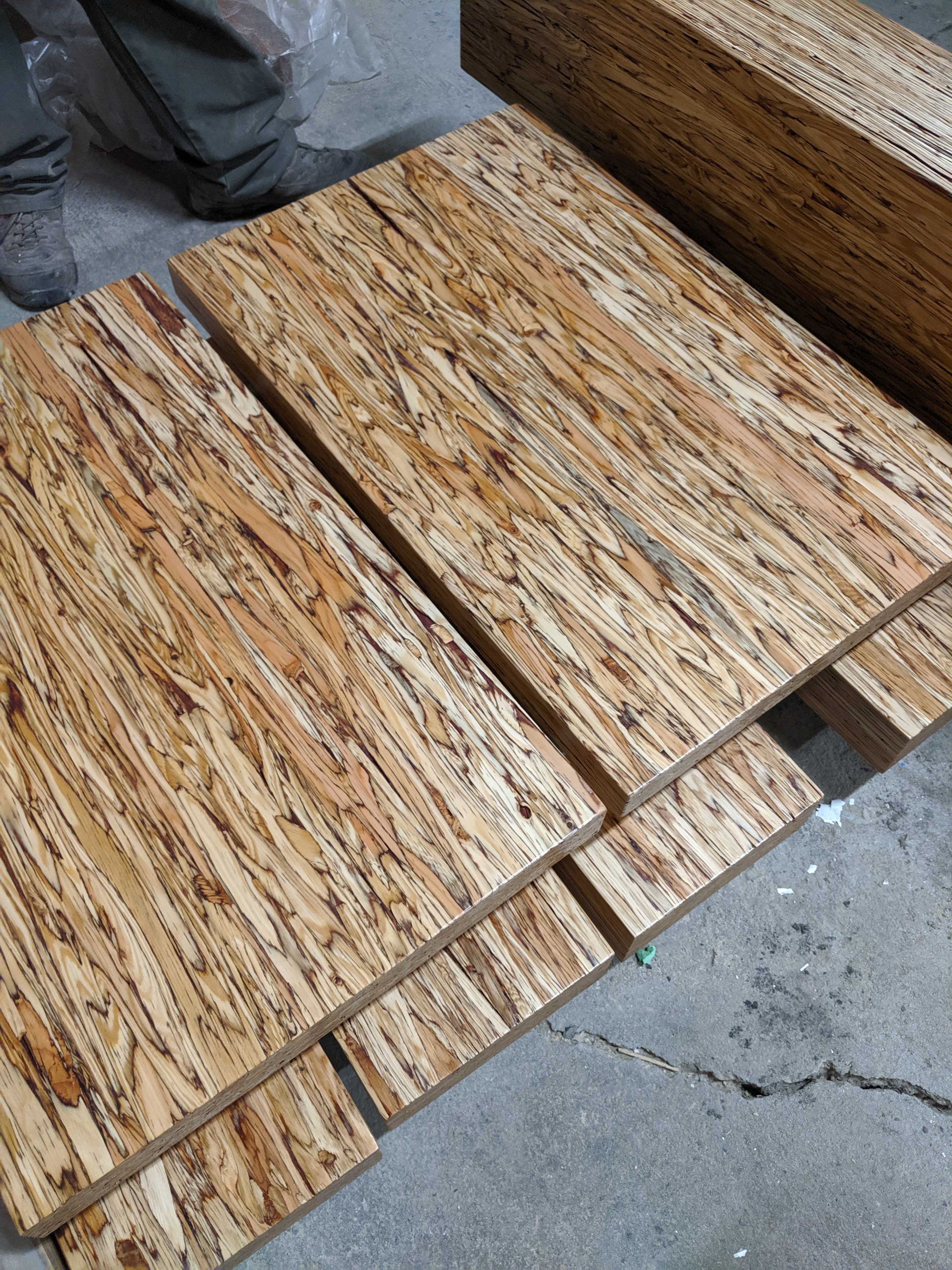 Stacked panels of textured, light-brown wood with distinct grain patterns on a concrete floor next to a person's legs.