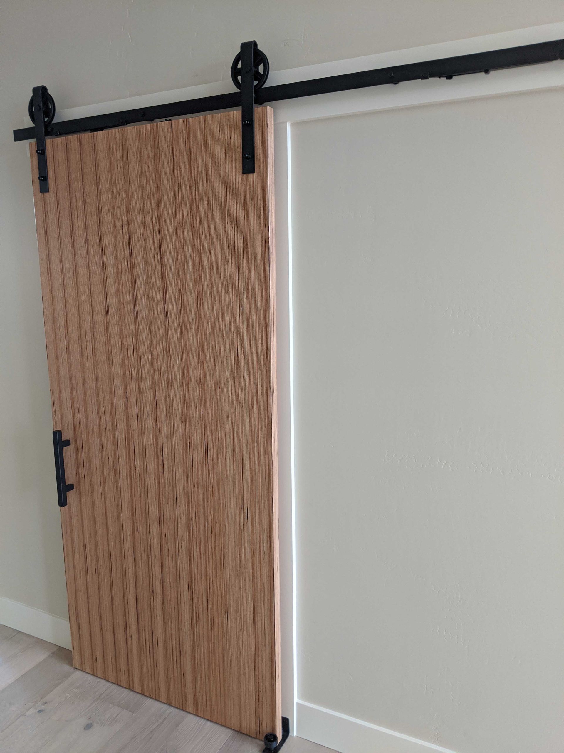 A light wood slatted barn door with black hardware, installed on a metal track over a white wall.