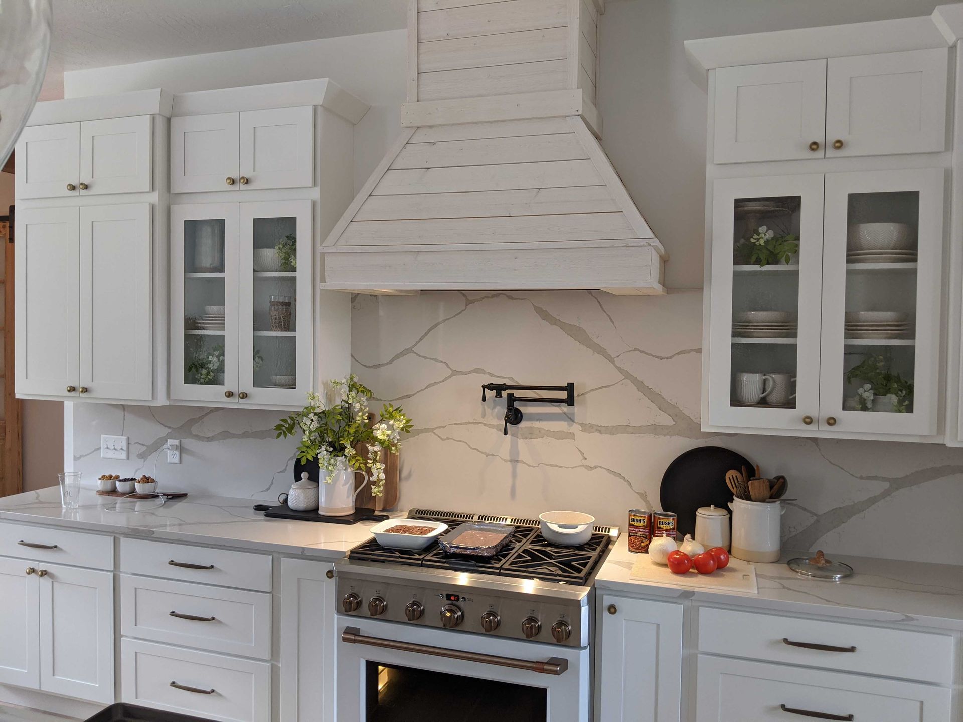 White farmhouse kitchen with glass-front cabinets, a rustic wood hood, marble-style backsplash, and stainless range.