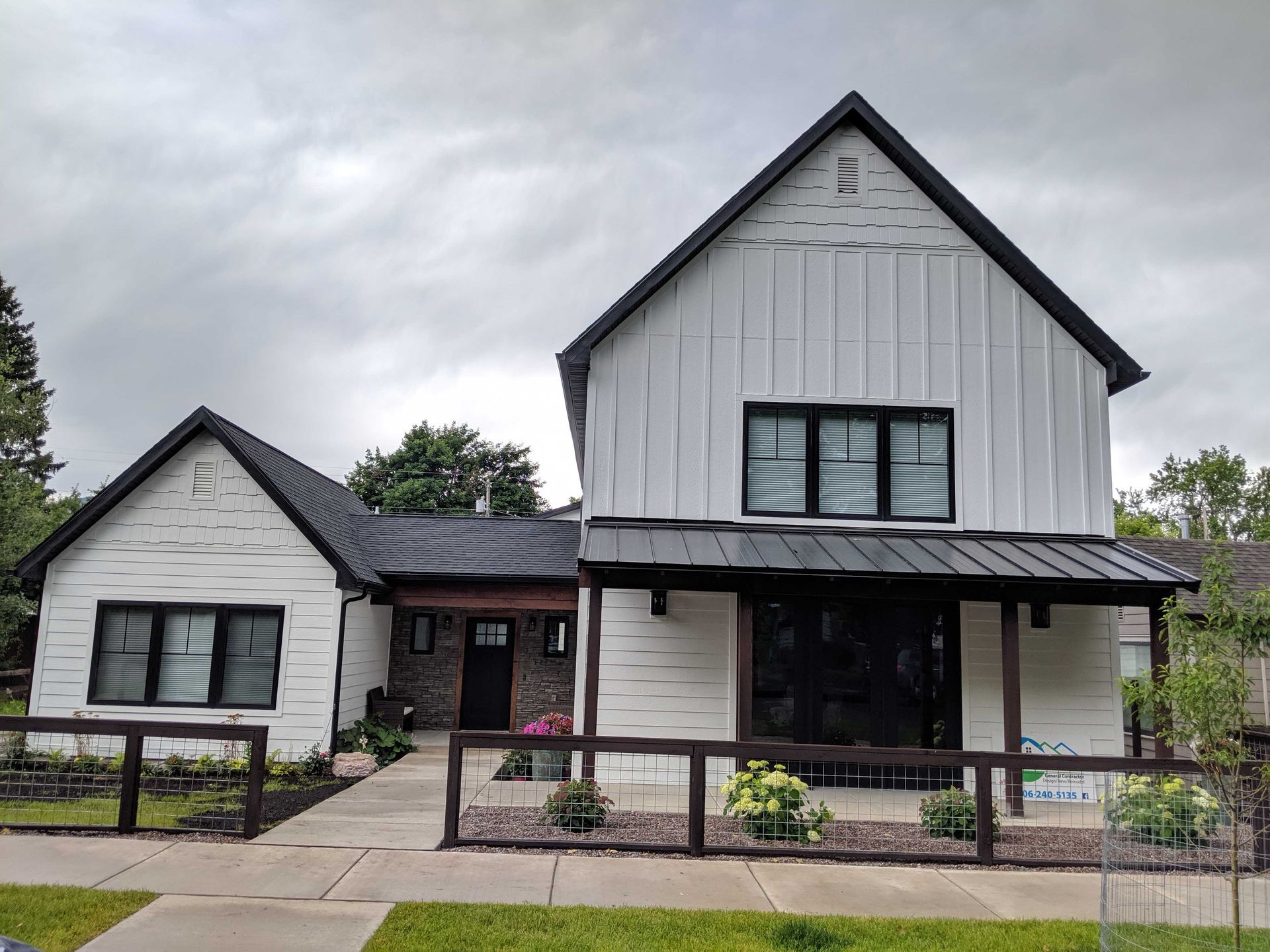 A modern white farmhouse with black trim, a gabled roof, and a front patio enclosed by a dark wood fence under a cloudy sky.