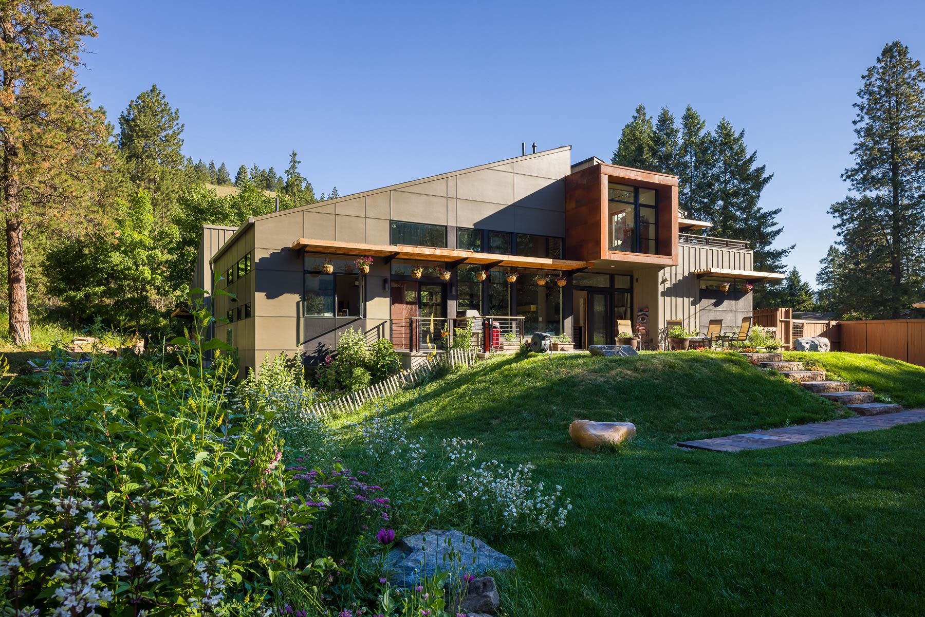 A modern house with a sloping roof and a wooden cantilevered box feature, set on a lush green lawn surrounded by trees.