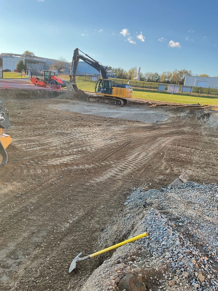 Construction site with a yellow excavator working on dirt ground, a red roller, and a shovel in the foreground.