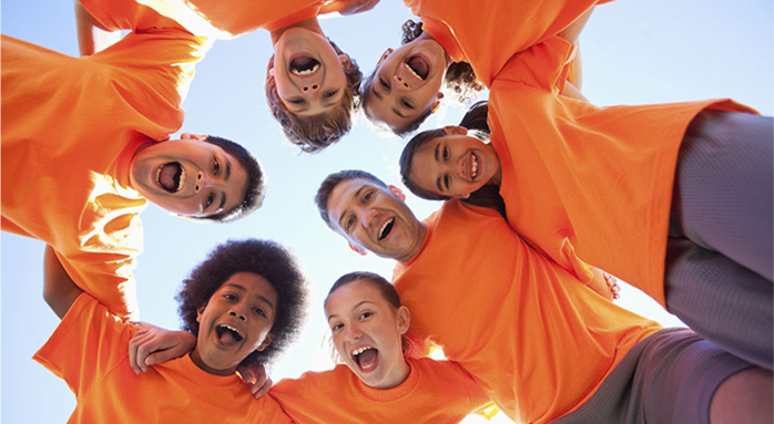 Friends in orange shirts huddle in a circle, smiling and looking down at the camera outdoors.