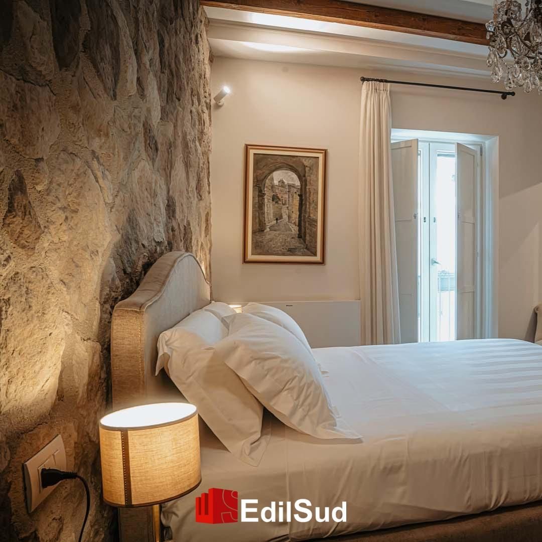Accogliente camera da letto con parete in pietra, letto bianco, lampada da comodino e quadro incorniciato vicino a una finestra luminosa.