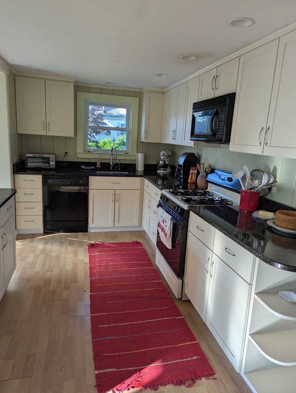 Kitchen with white cabinets, black appliances, red rug, and a window overlooking greenery.