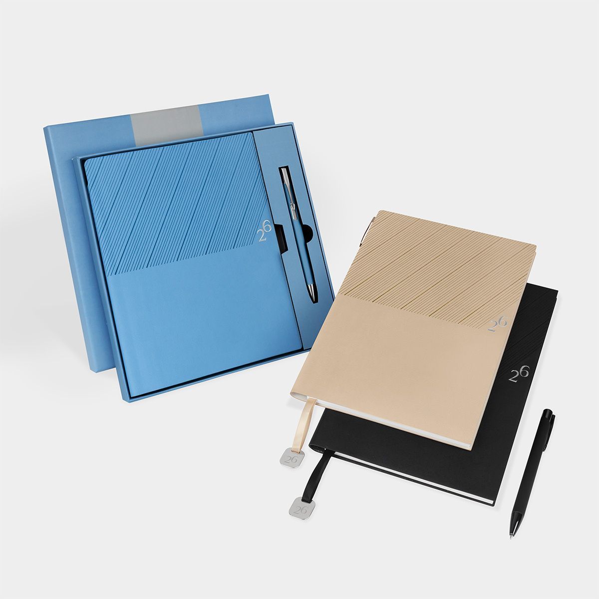 Tres cuadernos, uno azul con estuche, uno beige y uno negro, cada uno con un bolígrafo a juego sobre un fondo liso.