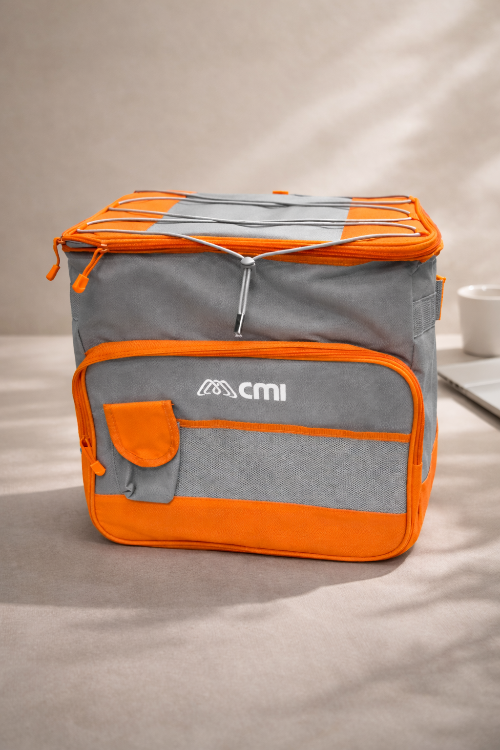 Una bolsa térmica gris y naranja con bolsillo frontal y cierre elástico en la parte superior, colocada sobre una superficie de color claro.