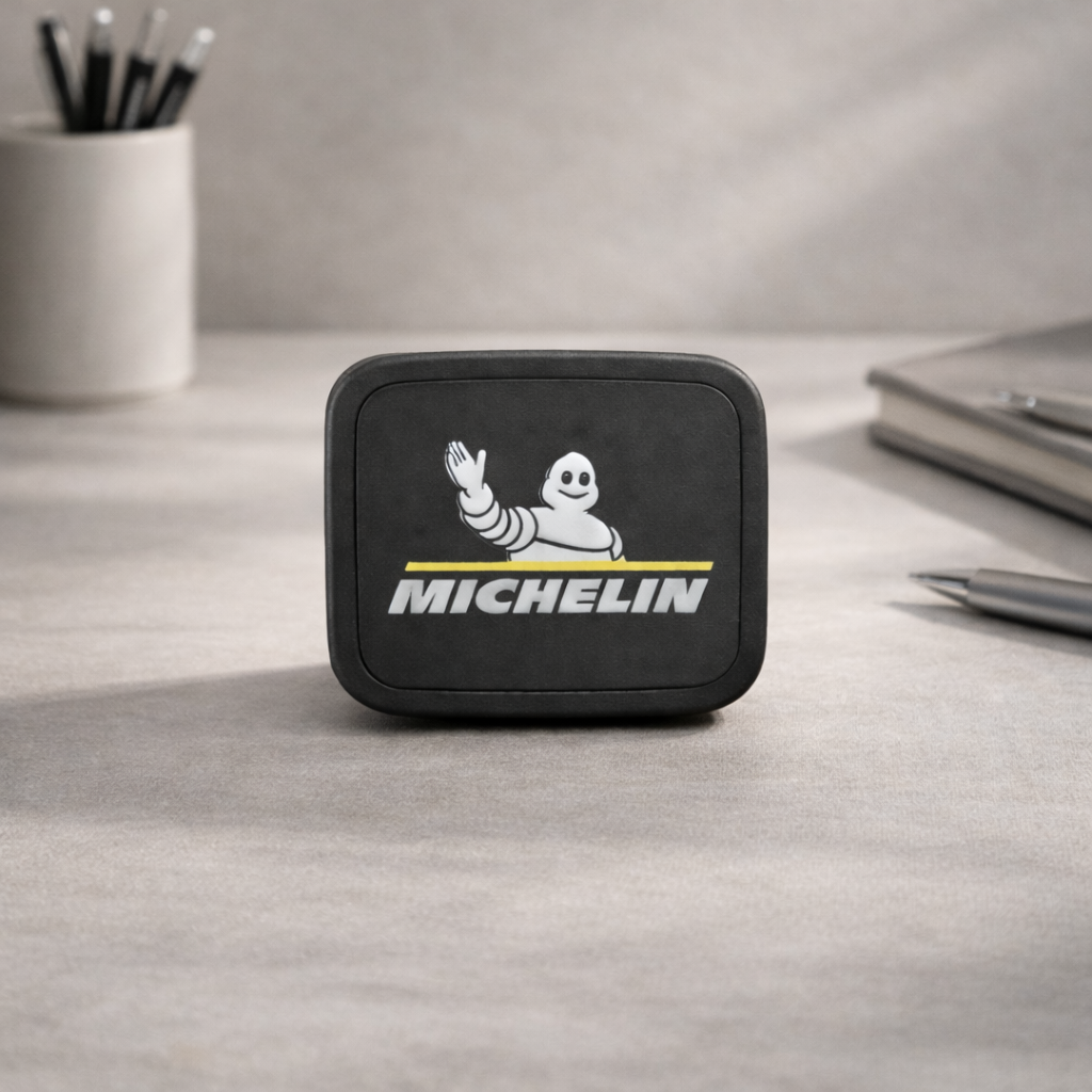Un objeto rectangular de color negro con la marca Michelin reposa sobre un escritorio gris, con un portalápices y un bolígrafo cerca.