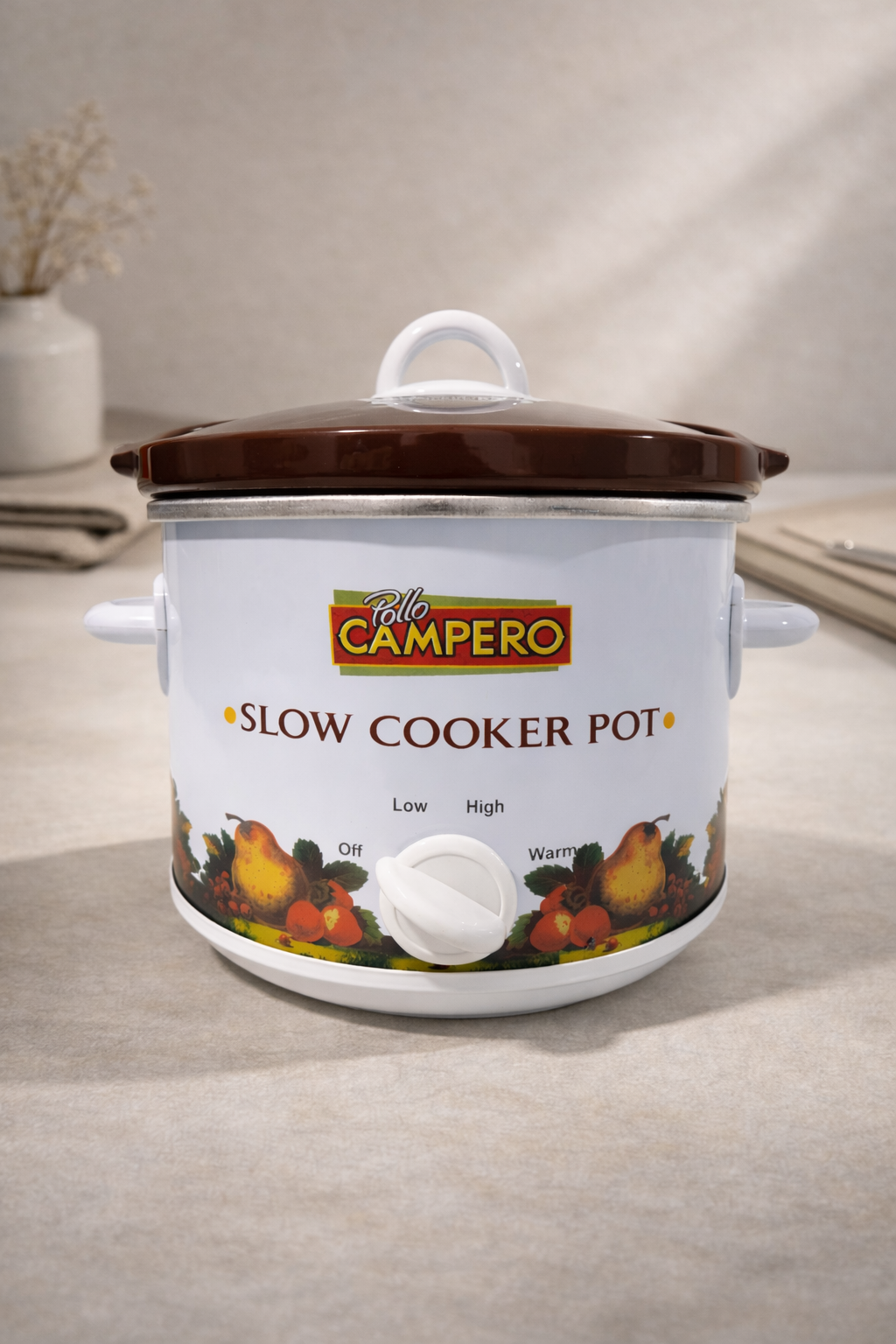 Una olla de cocción lenta Campero blanca con tapa marrón e ilustración de frutas reposa sobre una superficie neutra en una habitación bien iluminada.