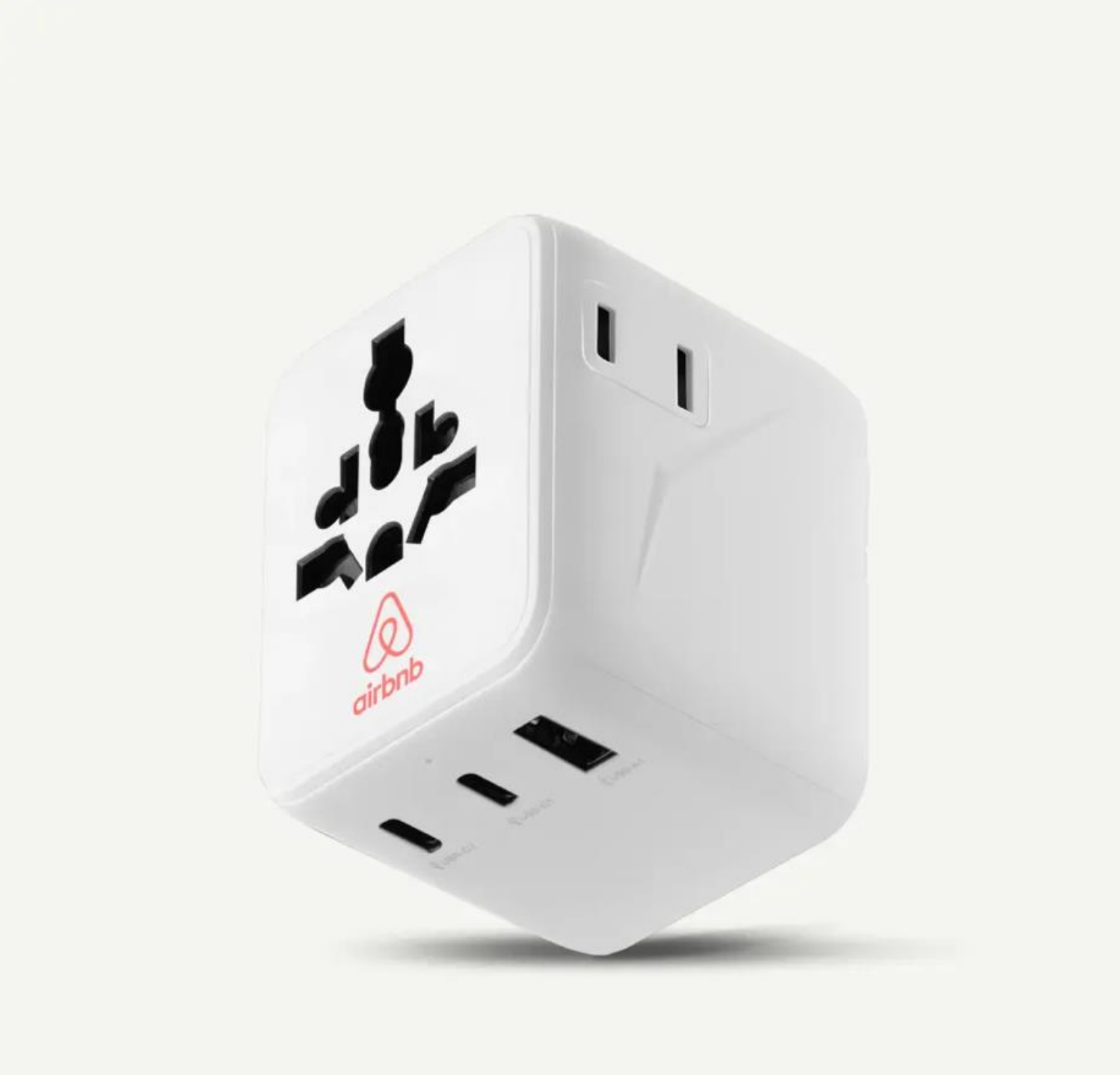 Un adaptador de viaje universal blanco con forma de cubo, con el logotipo de Airbnb y múltiples puertos USB y tomas de corriente internacionales.