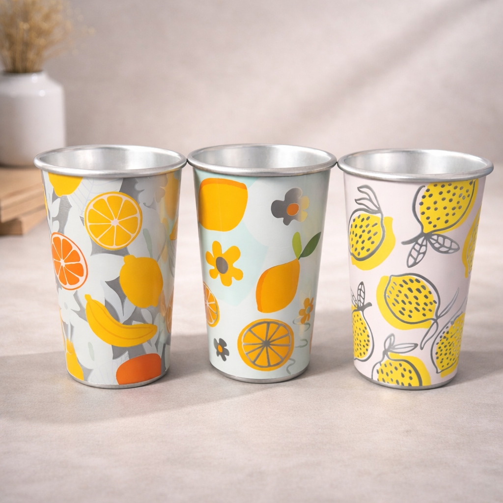 Tres vasos metálicos con diseños vibrantes de frutas, como limones, naranjas y plátanos, colocados sobre una superficie neutra.