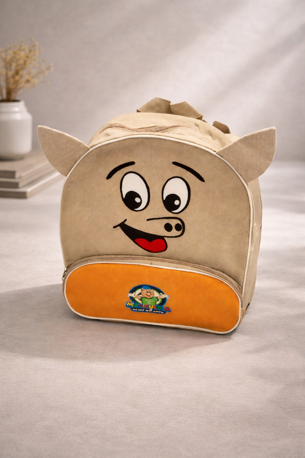 Una mochila infantil de tela color canela con forma de cerdito, con una carita sonriente y un bolsillo frontal naranja con un logotipo circular.