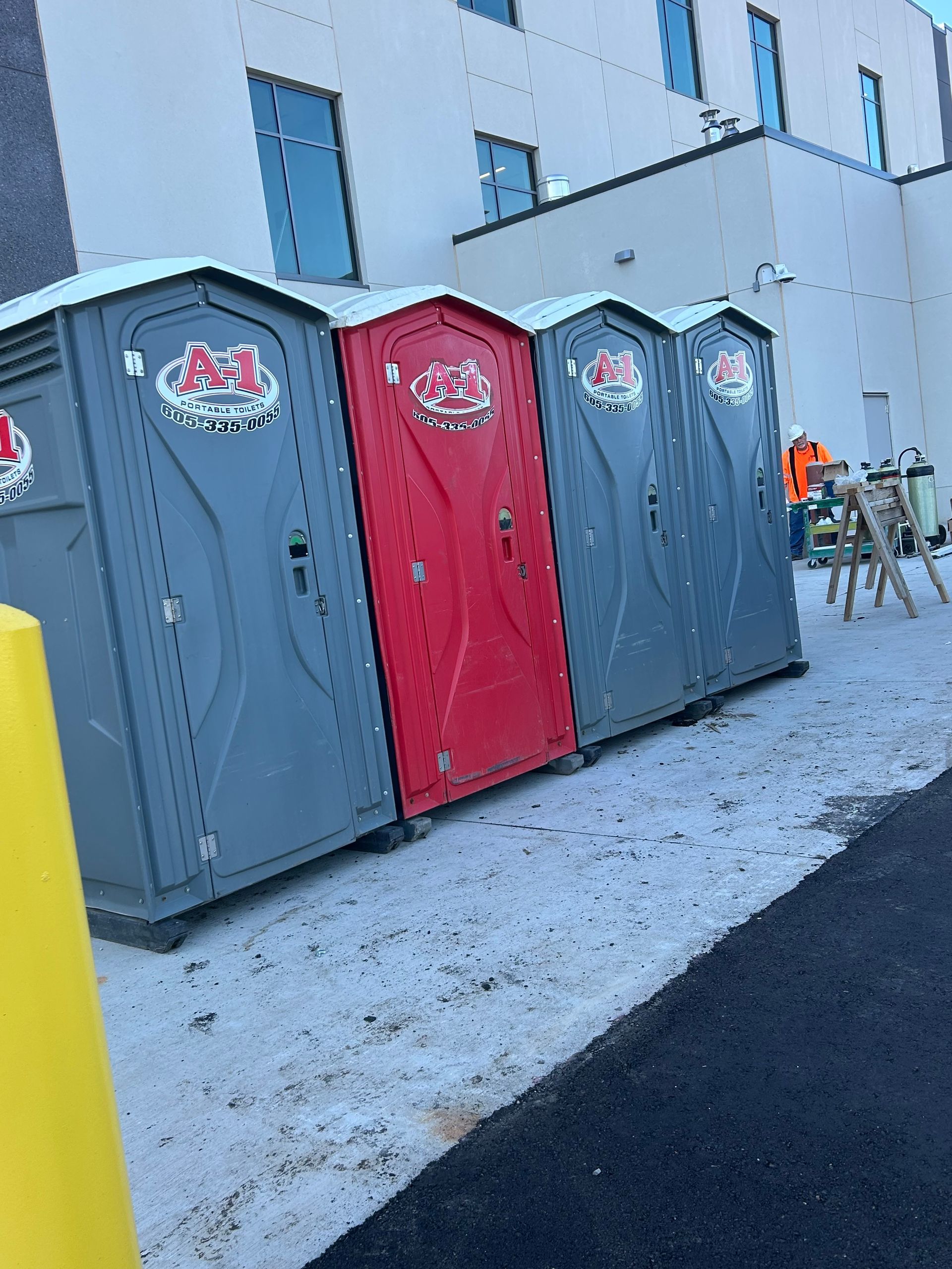 Three portable toilets, two gray and one red, beside a building on a snowy sidewalk.
