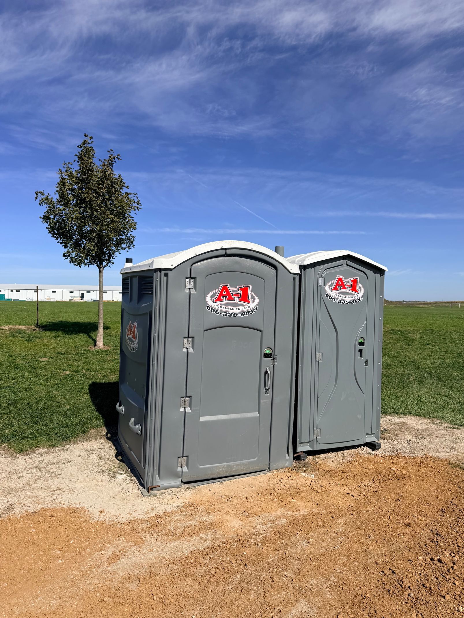 Two gray portable toilets on a gravel path in an open field under a blue sky