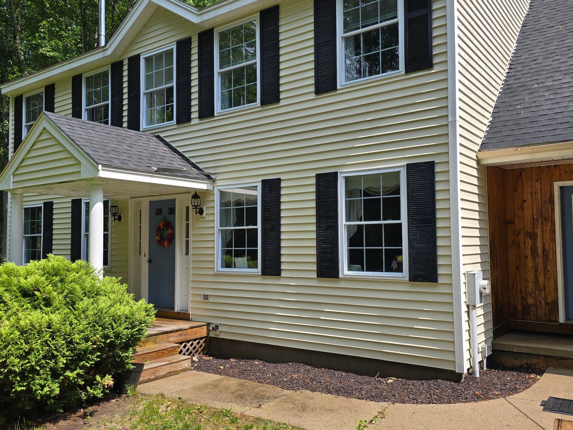 Suburban two-story house with beige siding, white trim, front porch, and a shrub by the entrance